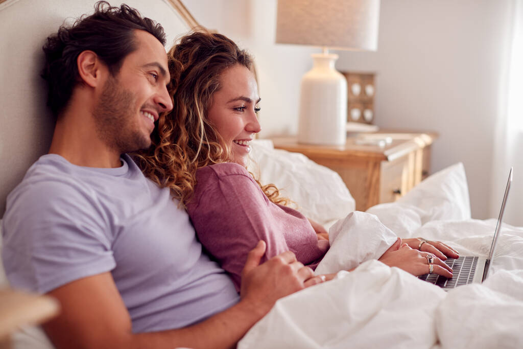 Loving coppia indossa pigiama sdraiato a letto a casa guardando computer portatile insieme - Foto, immagini