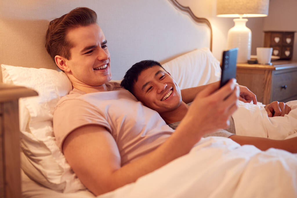 Αγαπώντας το ίδιο φύλο αρσενικό ζευγάρι που βρίσκεται στο κρεβάτι στο σπίτι κοιτάζοντας τα μέσα κοινωνικής δικτύωσης στο κινητό τηλέφωνο - Φωτογραφία, εικόνα