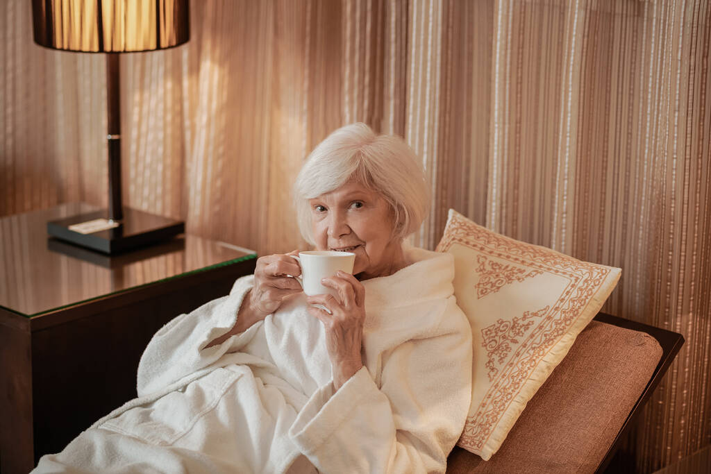 Een oudere grijze vrouw die in bed ligt en 's ochtends koffie drinkt. - Foto, afbeelding