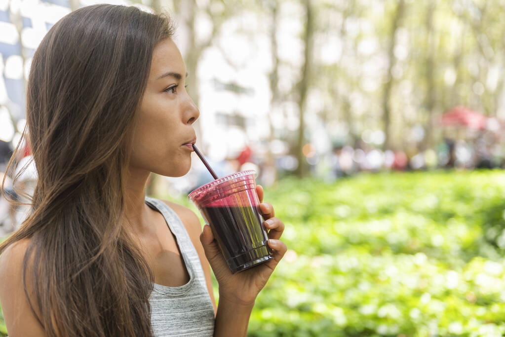 Woman drinking beet juice smoothie walking in city park. Girl eating detox food from online delivery service. Young asian biracial woman young adult living healthy lifestyle - Foto, imagen