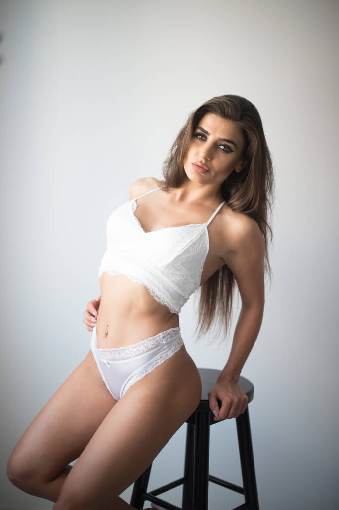 Attractive sexy brunette in white lingerie  posing provocatively, in front of a wall ,studio shot. Portrait of sensual woman with long hair, white bikini and bra in classic boudoir scene in studio - Photo, Image