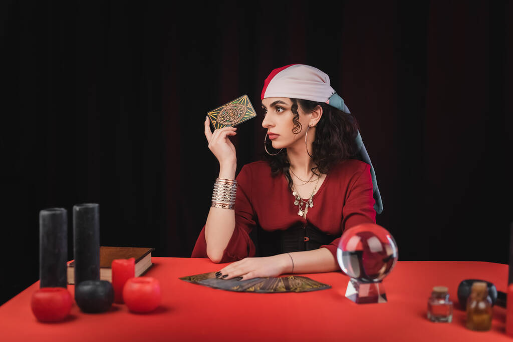 KYIV, UKRAINE - FEBRUARY 23, 2022: Young gypsy fortune teller holding tarot near witchcraft supplies on table isolated on black  - Fotografie, Obrázek