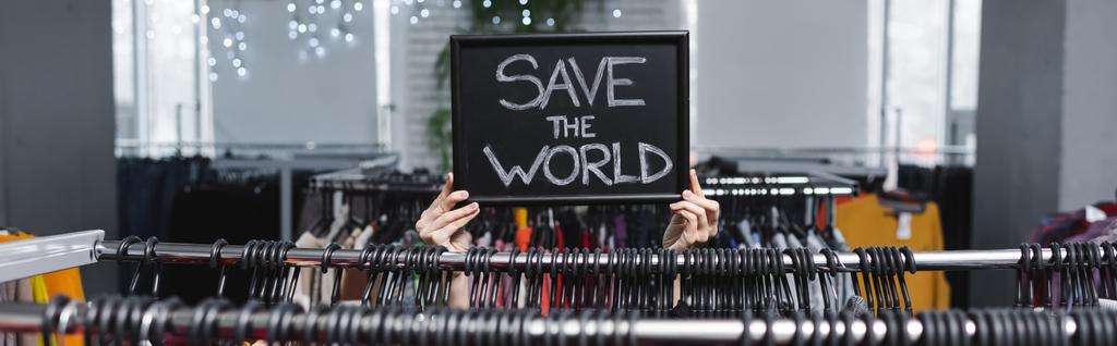 Cropped view of saleswoman holding board with save the world lettering near hangers in vintage shop, banner  - Foto, afbeelding