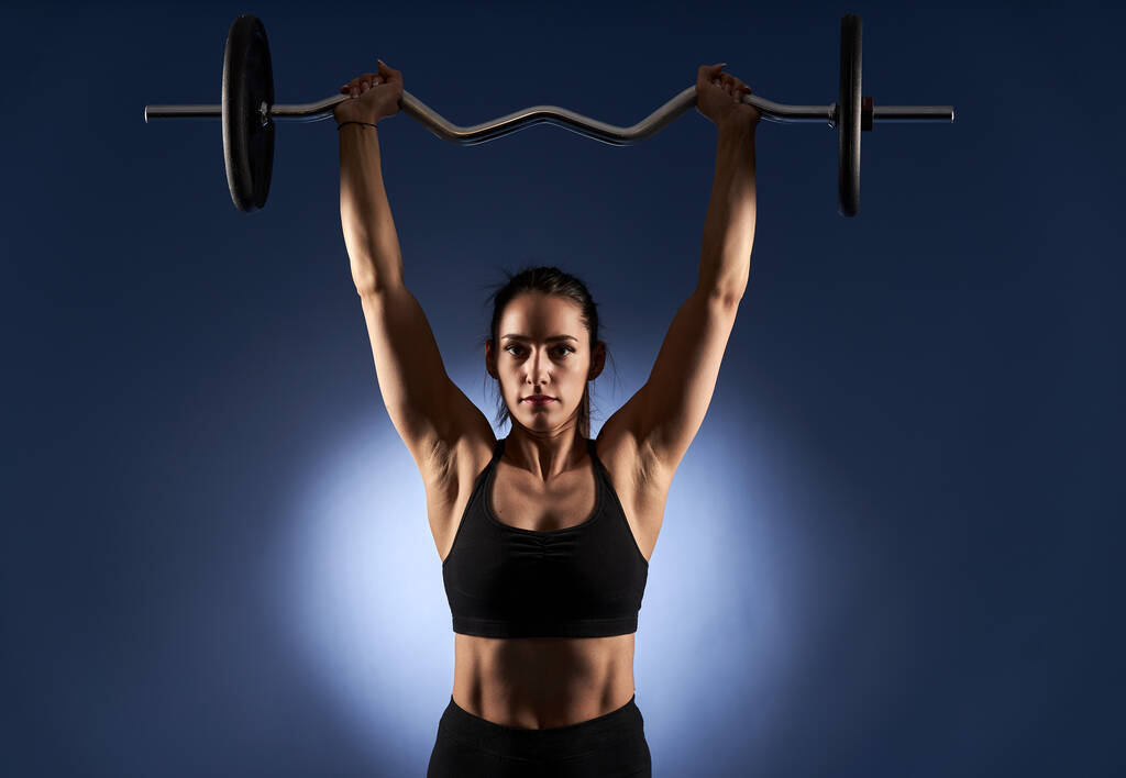 Jonge atletische vrouw doet schouder training met lange halter op blauwe achtergrond - Foto, afbeelding