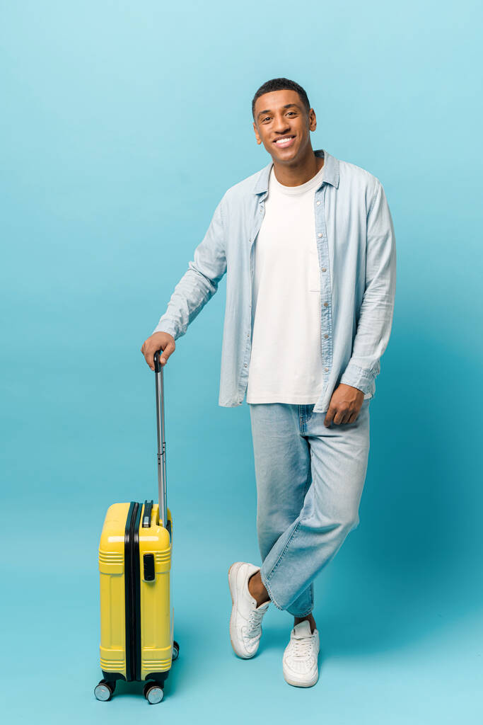 Teljes hossz fotó mosolygós fiatal afro-amerikai srác alkalmi farmer viselet álló sárga bőrönd - Fotó, kép