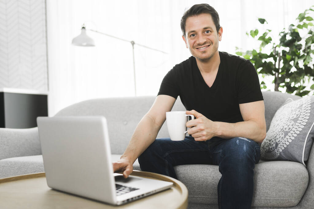 European man holding coffee while working on his laptop in bright living room. - Фото, изображение