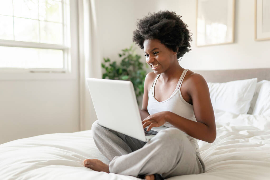 junge schwarze Frau mit Laptop im hellen Schlafzimmer - Foto, Bild