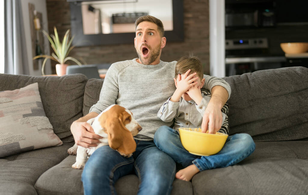 Vater oder Pate mit Basset-Hund amüsieren sich auf dem Wohnzimmer Zu Hause sehen Filme mit Popcorn wie Angst aus - Foto, Bild