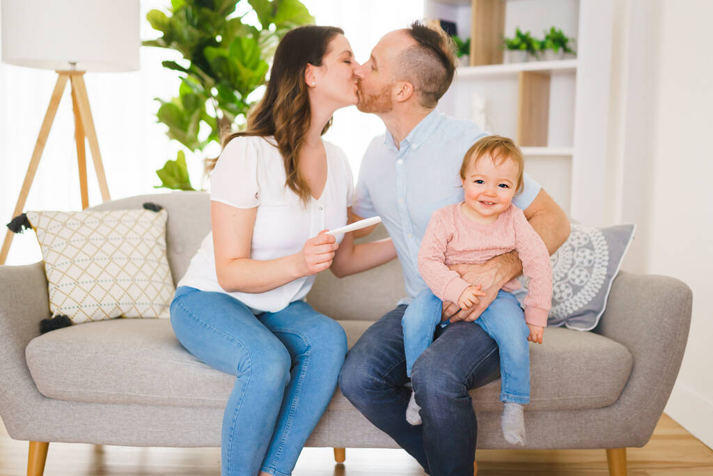 自宅のソファに座って興奮した妻と妊娠検査に満足している家族. - 写真・画像