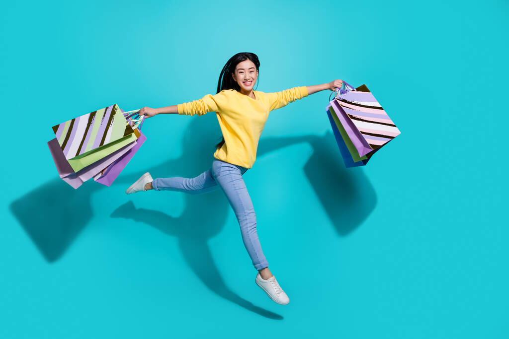 Full length photo of funny excited lady dressed yellow sweater jumping high holding shoppers isolated turquoise color background - Zdjęcie, obraz