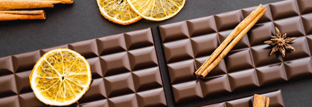 Siyah arka plandaki çikolataların üzerinde tarçın ve kuru turuncu çubukların üst görünümü, afiş  - Fotoğraf, Görsel