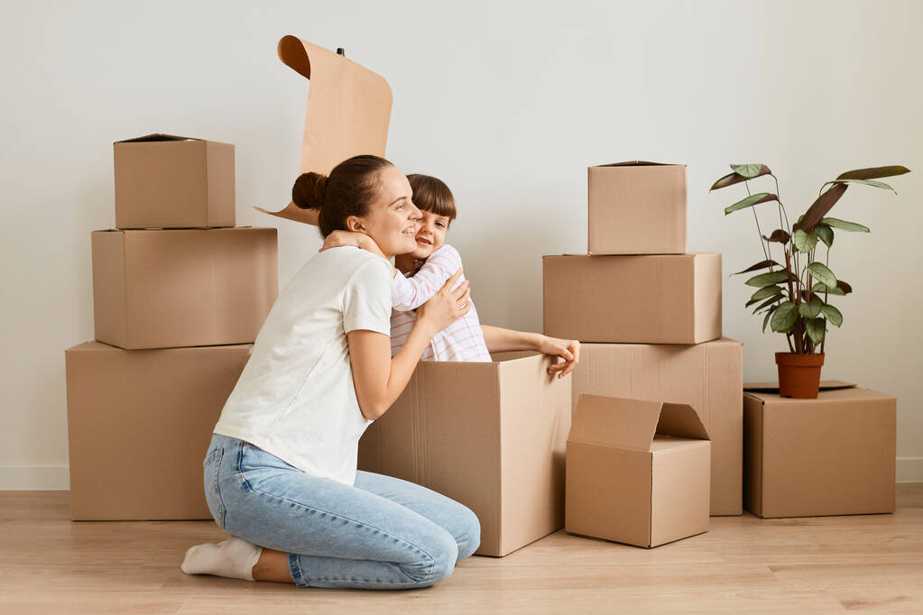 Image of little girl with her mother sitting in handmade boat from cardboard boxes and hugging, happy child playing with mommy during moving in a new flat. - 写真・画像