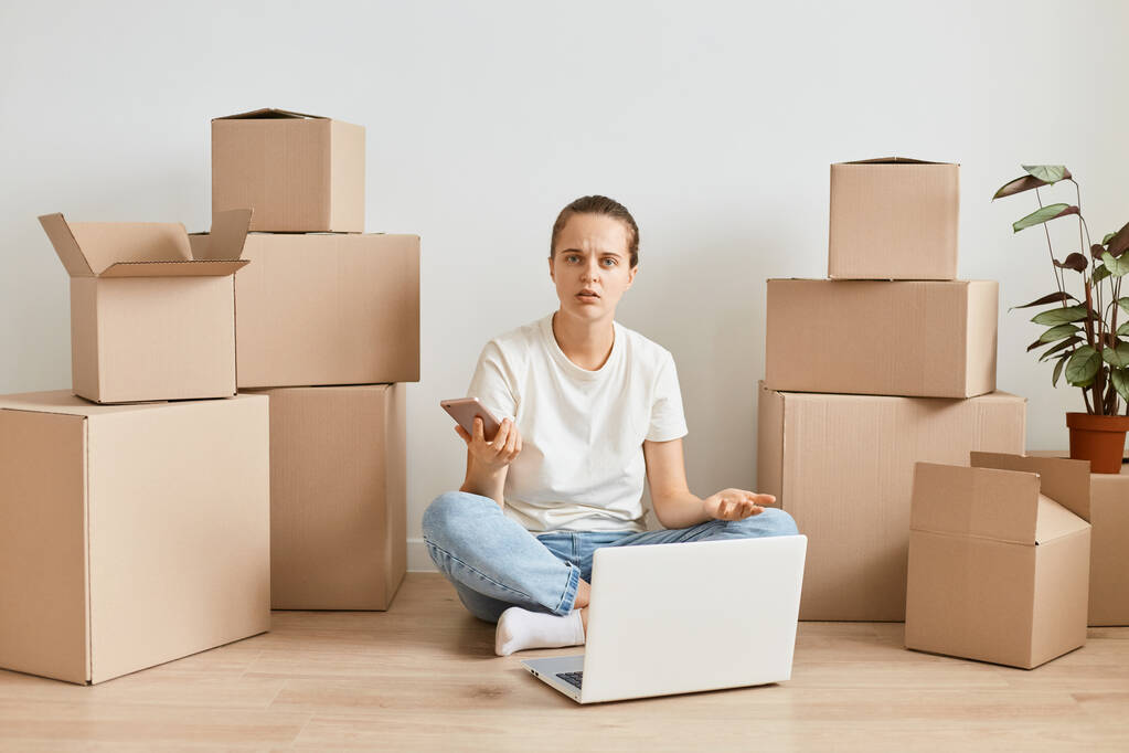 Portrait of confused young adult female wearing jeans and white t shirt sitting on floor near carton boxes and holding phone, looking at camera with puzzlement, relocating. - Foto, Imagem