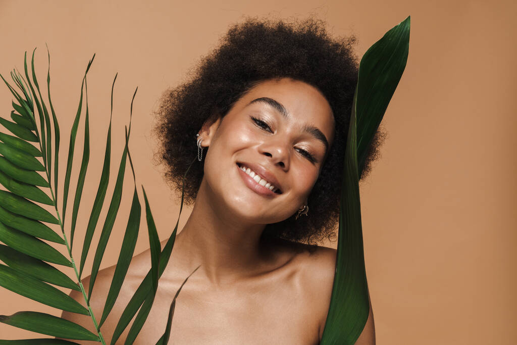 Black shirtless woman smiling while posing with green leaves isolated over beige background - Fotografie, Obrázek
