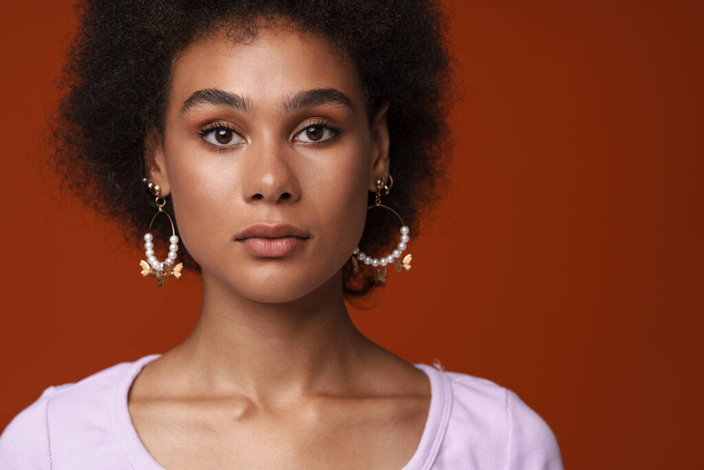 Black young woman in earrings posing and looking at camera isolated over brown background - Photo, Image