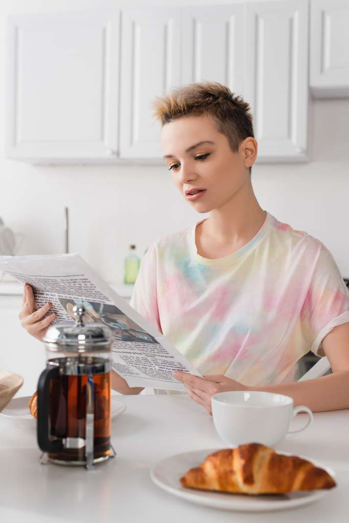 pessoa bigender com cabelo curto leitura jornal da manhã perto bule e croissants borrados - Foto, Imagem