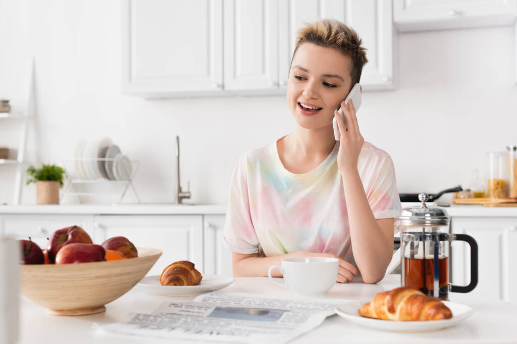 usmívající se pangender osoba mluví na mobilním telefonu v blízkosti croissanty, konvice a čerstvá jablka - Fotografie, Obrázek