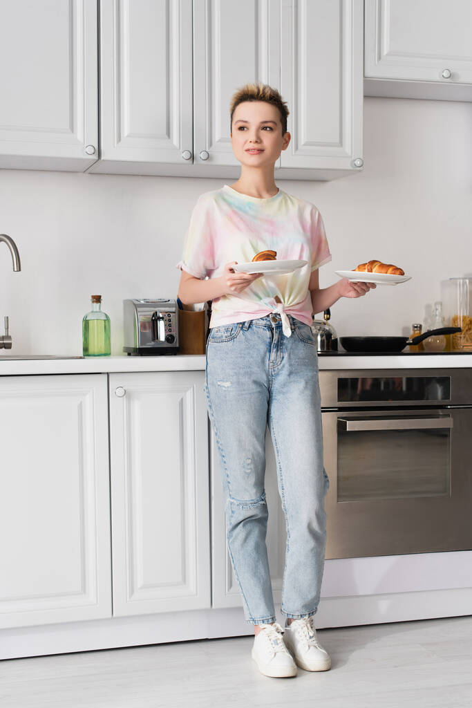 full length of positive pangender person in jeans holding tasty croissants in kitchen - Foto, immagini