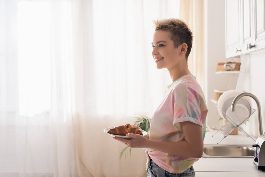 вид сбоку счастливого бигендера с короткими волосами, держащими тарелку с круассаном на кухне - Фото, изображение