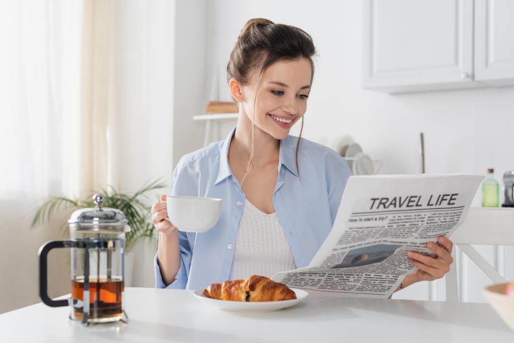 smiling woman with cup of tea reading travel life newspaper near delicious croissant - Fotoğraf, Görsel
