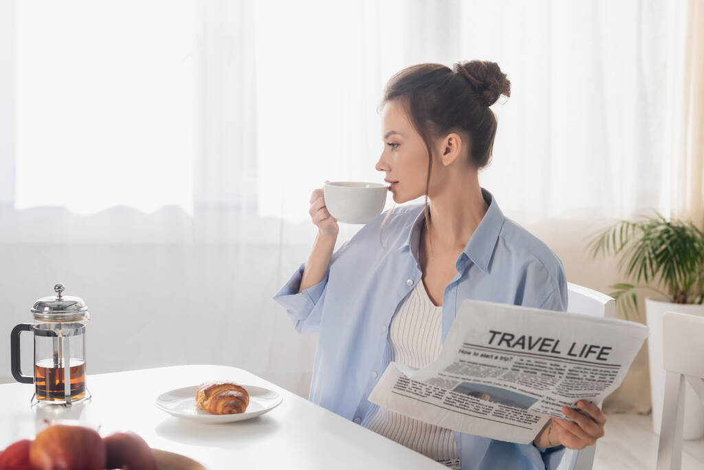 young woman with travel life newspaper drinking tea near tasty croissant - Foto, afbeelding