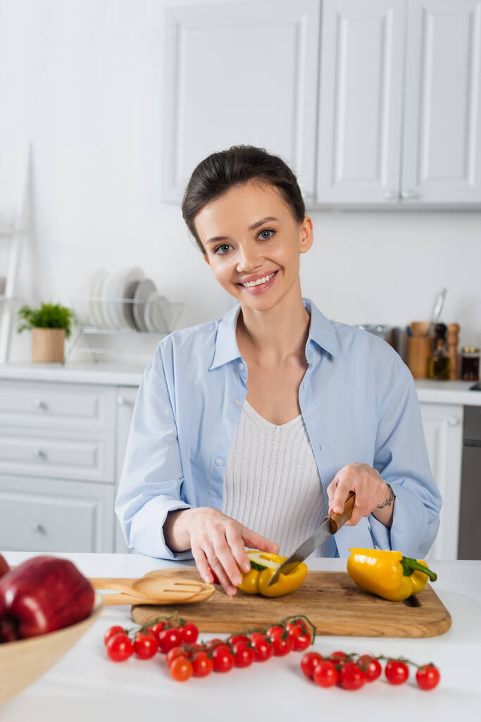 радісна жінка, що ріже болгарський перець біля свіжих вишневих помідорів на кухні
 - Фото, зображення