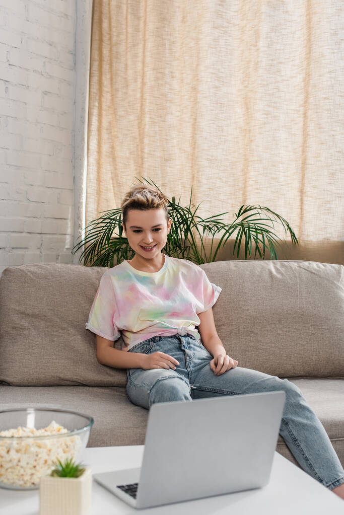 junge und glückliche pansexuelle Person mit kurzen Haaren, die auf der Couch sitzt und sich Filme auf dem Laptop ansieht - Foto, Bild