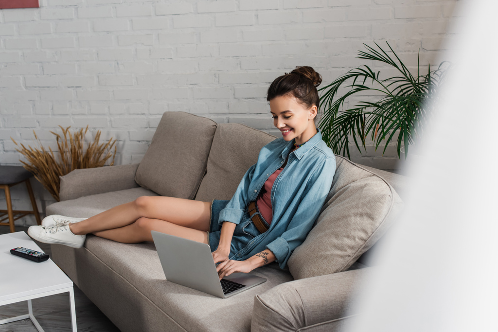 full length of cheerful woman sitting on couch and typing on laptop on blurred foreground - Fotoğraf, Görsel