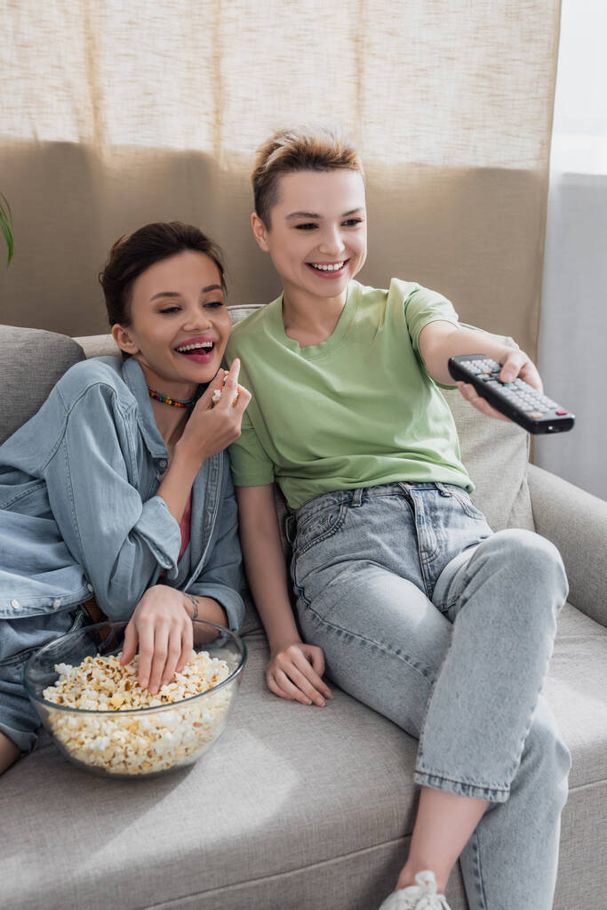 cheerful bigender person clicking tv channels near lover with bowl of popcorn - Foto, imagen