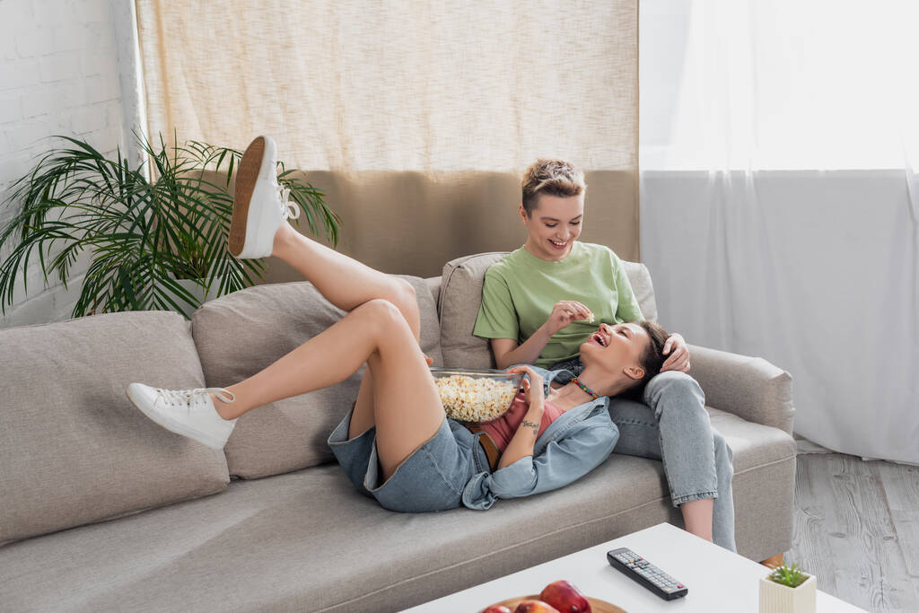 bigender person feeding cheerful partner with popcorn on sofa in living room - Foto, immagini