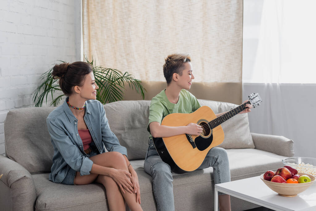 młody muzyk bigender gra na gitarze akustycznej w pobliżu partnera na kanapie w salonie - Zdjęcie, obraz