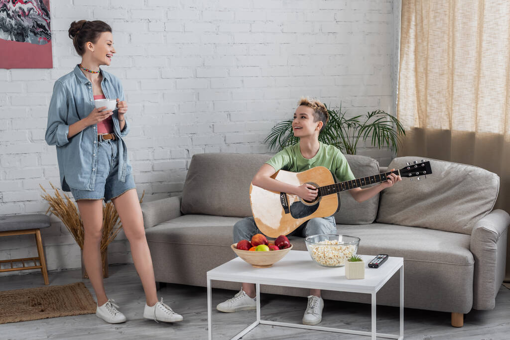 bigender muzyk gra na gitarze na kanapie obok uśmiechniętego partnera stojącego z filiżanką herbaty - Zdjęcie, obraz
