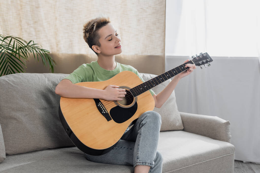 smiling bigender person with short hair sitting on couch and playing acoustic guitar - Zdjęcie, obraz