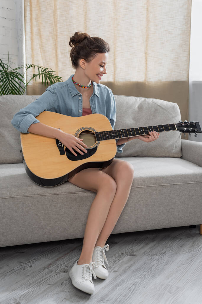 pełny widok długości wesoły kobieta siedzi na kanapie i gra na gitarze akustycznej - Zdjęcie, obraz