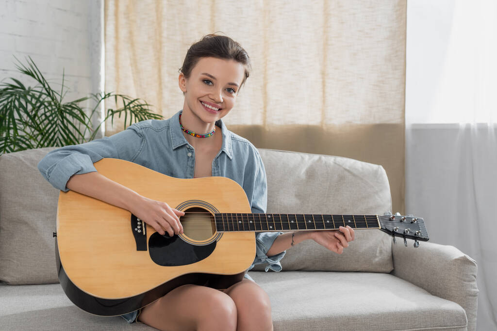 brunette woman smiling at camera while playing acoustic guitar on couch in living room - Photo, Image
