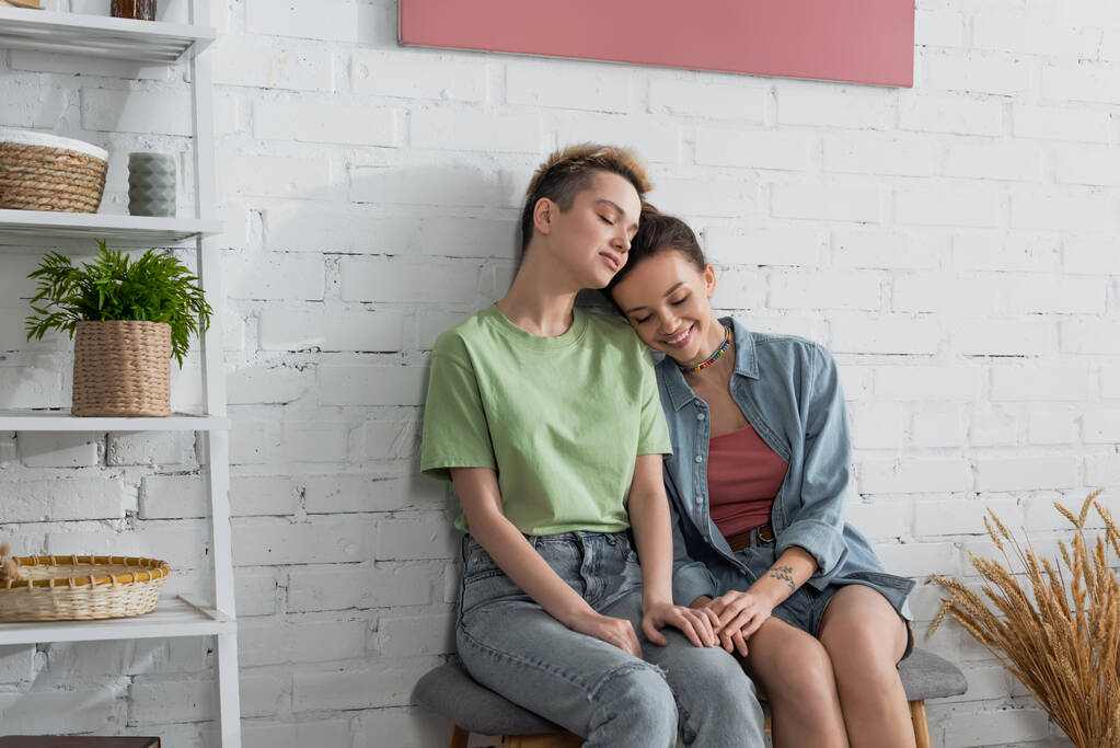 young and happy pangender couple with closed eyes sitting on ottoman near white brick wall - Foto, Bild