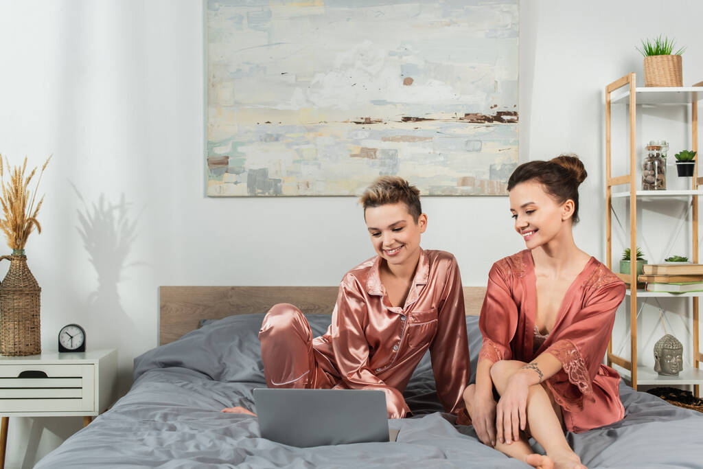 happy pangender lovers in satin pajamas and robe watching film on laptop in bedroom - Foto, imagen