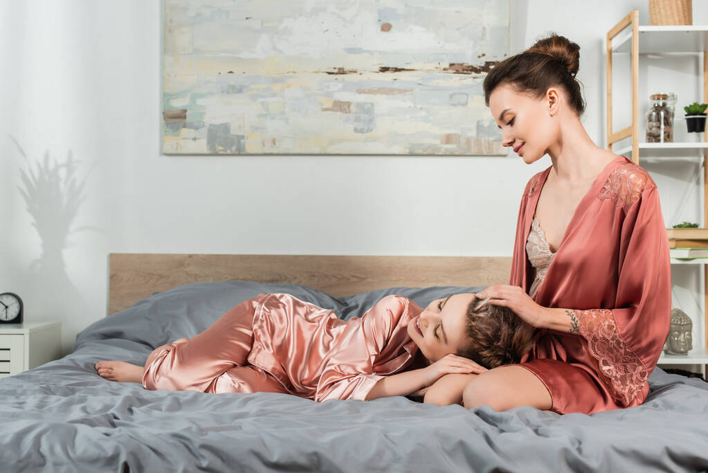 sonriente pansexual persona tocando pelo de amante relajándose en cama en satén pijama - Foto, imagen