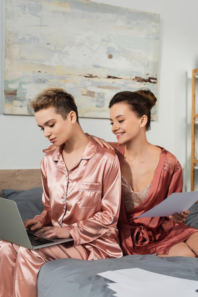 Mutlu panseksüel kişi yatak odasında dizüstü bilgisayarda yazan partnerinin yanında belge tutuyor. - Fotoğraf, Görsel