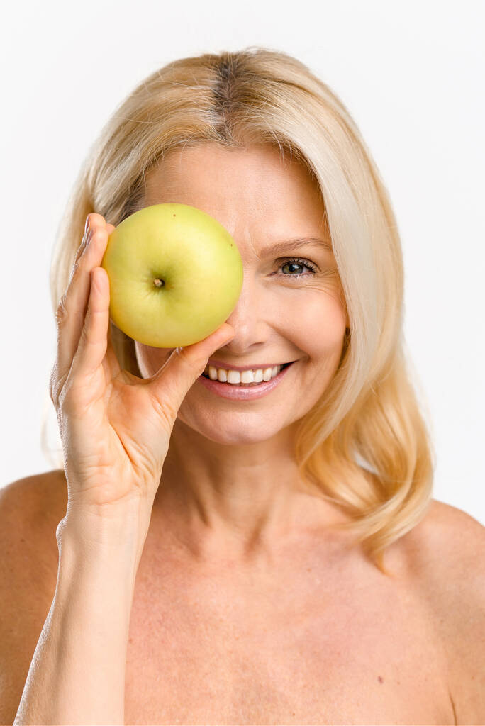 Cheerful joyful mature 50s woman covered one eye with fresh green apple and laughing. Middle-aged model with naked shoulders isolated on white - Foto, Imagem