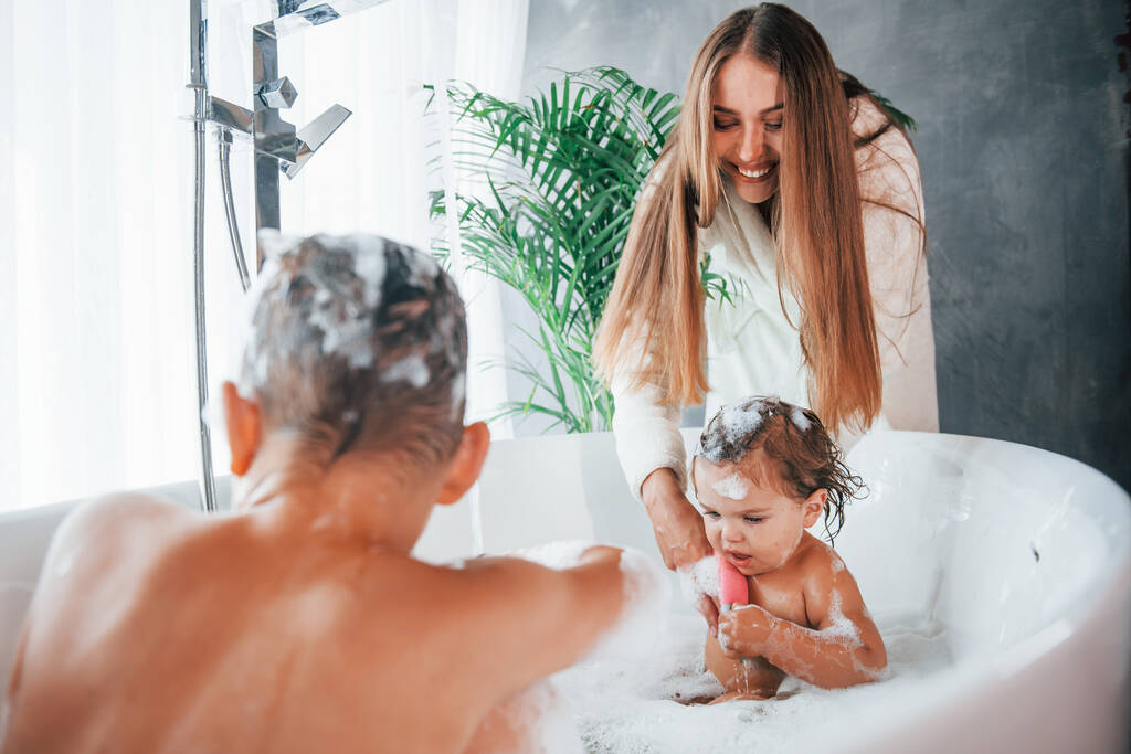 Bavte se. Mladá matka pomáhá svému synovi a dceři. Dvě děti se myjí ve vaně. - Fotografie, Obrázek