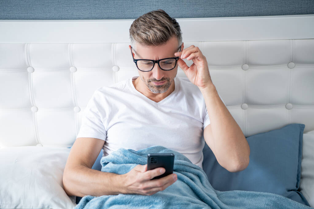 homem maduro na cama conversando no telefone vestindo óculos - Foto, Imagem