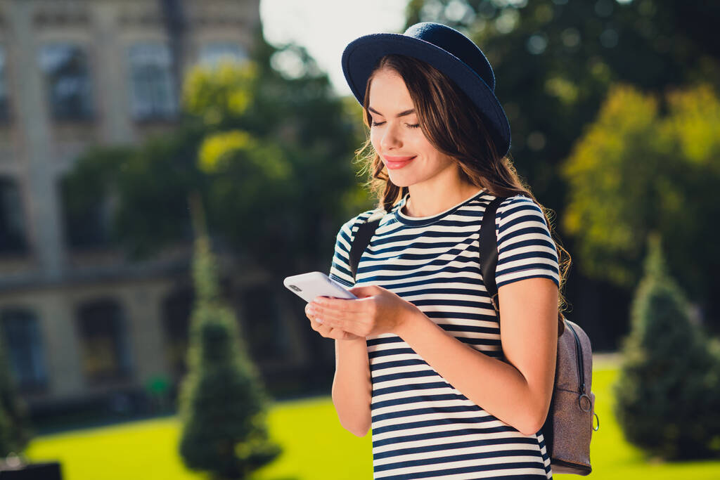 Photo of sweet cute young woman dressed striped outfit cap backpack writing message modern device smiling outside landscape - Фото, изображение