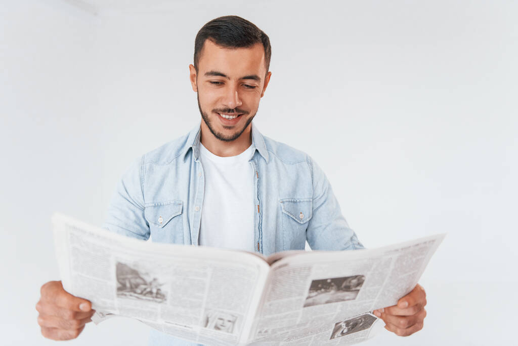 Διαβάζει εφημερίδα. Νεαρός όμορφος άντρας στέκεται μέσα σε λευκό φόντο. - Φωτογραφία, εικόνα