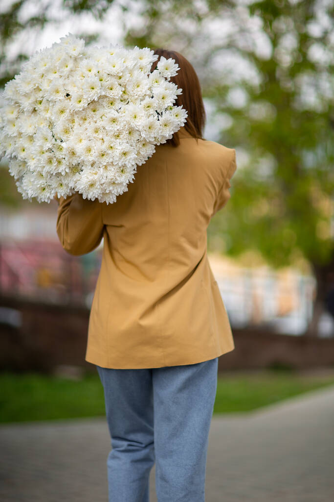 Young brunette female in modern beige outfit with bunch of chryzantem flowers walking in sunny european city street. Back view. Aesthetics. Beautiful girl with big daisies bouquet in field - Foto, Imagen