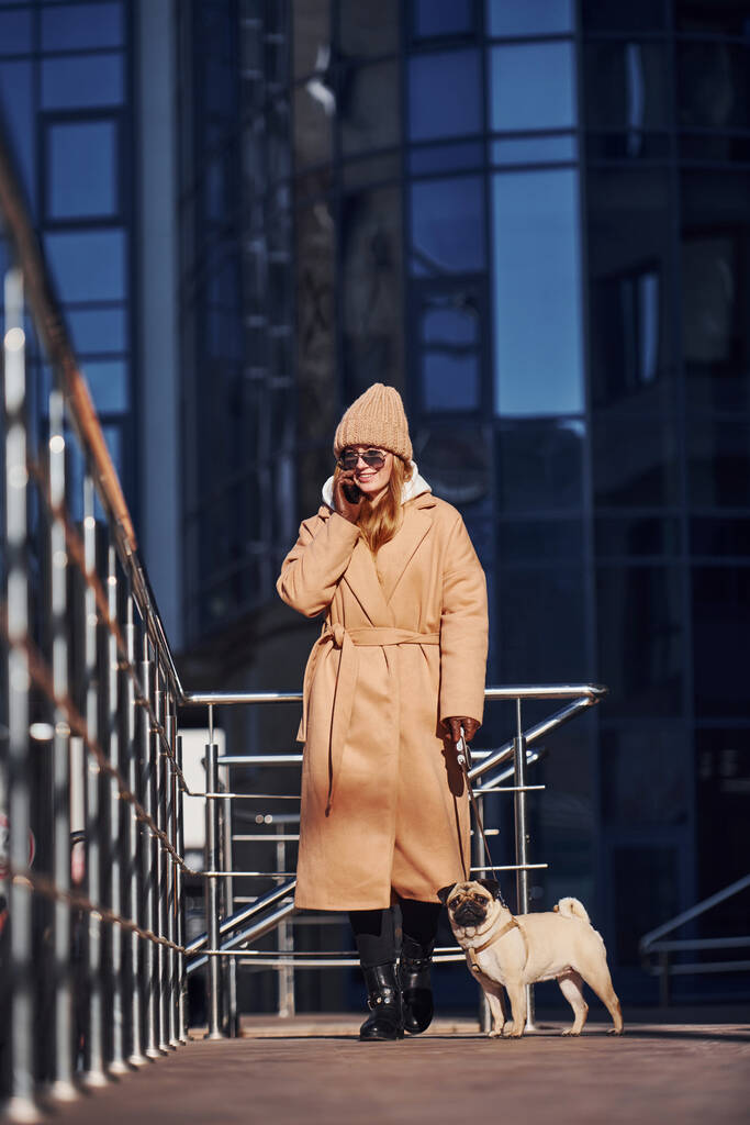 Жінка в теплому одязі гуляє зі своєю маленькою мопсовою собакою біля бізнес-будівлі, яка на фоні і розмовляє по телефону
. - Фото, зображення