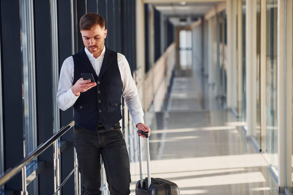 Junger männlicher Passagier in eleganter Festkleidung steht mit Gepäck und Telefon in der Hand in der Flughafenhalle. - Foto, Bild