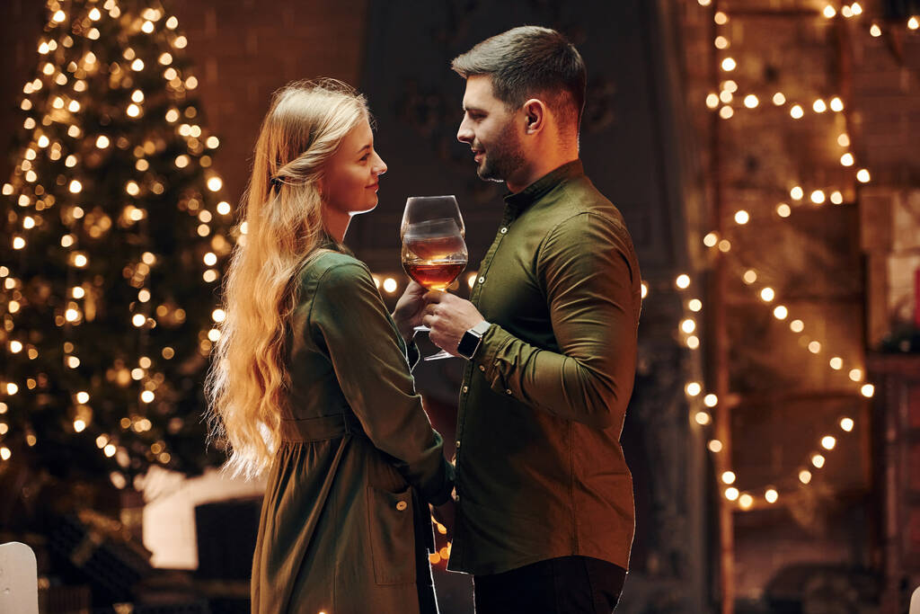 De pie y sosteniendo grandes vasos con vino. Joven pareja encantadora tienen cena romántica en el interior juntos. - Foto, Imagen
