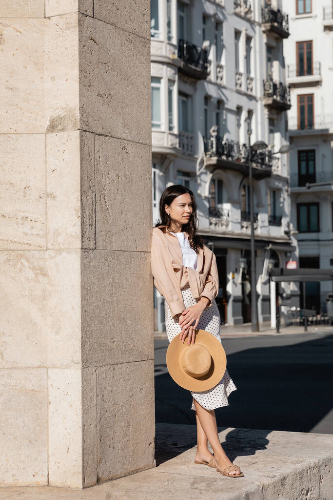 full length view of stylish woman with straw hat standing near wall and looking away - Foto, immagini