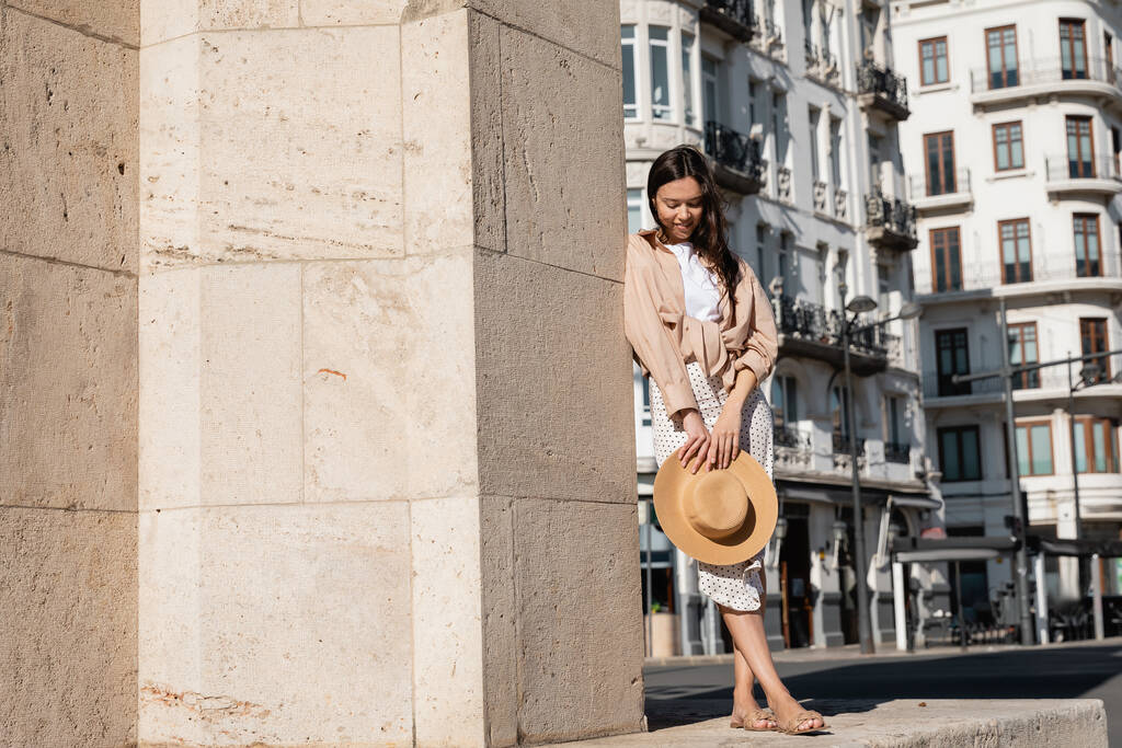 full length of stylish woman with straw hat near wall on urban street - Фото, изображение